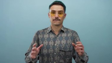 Mavi arka planda sarı gözlüklü genç adam şaşırmış görünüyor, elleri havada desenli bir gömleği vurguluyor, canlı ve anlamlı bir duygu ve tarz sahnesi yaratıyor..