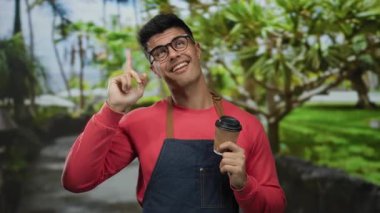 Gözlüklü, kırmızı ve önlüklü, elinde kahve fincanı tutan genç adam, gülümsüyor ve yemyeşil bir arka planda yukarı doğru bakıyor..