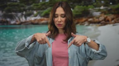 İspanyol kadın deniz kenarını işaret ediyor, kot ve pembe gömlek giyiyor, deniz manzaralı açık havada duruyor, sakin sahil ortamında kendine güven ve sükunet yayıyor..