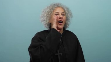 Dini cüppeli, kıvırcık gri saçlı yaşlı bir kadın, dışa kapalı mavi arka planda, anlamlı jestler ve yüz ifadesi yoluyla inancını ve duygularını ifade eden tutkulu bir şekilde konuşuyor.