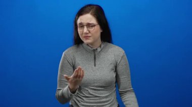 Stüdyo ortamında elleri göğsünde, mavi arka planda duygularını ifade eden, gri süveter ve gözlük takan, samimi ve içten duygular gösteren bir kadın..