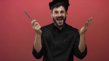 Sakallı bir adam aşçı kıyafeti giymiş elinde peynir rendesiyle kırmızı arka planda duruyor, neşeli bir mutfak havası yayıyor..