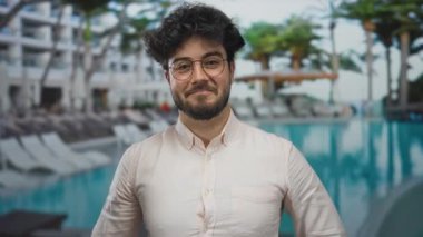 Sakallı genç İspanyol adam tatil köyünün havuzunda kendini parmaklarıyla göstererek lüks otel arka planına karşı kendine güvenen bir ifade sergiliyor..