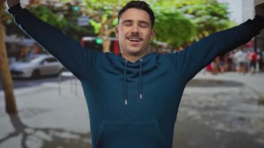 Telaşlı bir şehir caddesinde kollarını açmış gülümseyen genç İspanyol bir adam, yeşil kapüşonlu bir kapüşonlu giyiyor, şehir zemininde bir neşe ve özgürlük anı yakalıyor..