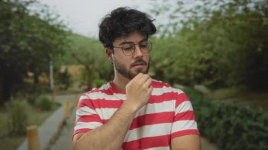 Sakallı ve gözlüklü genç bir adam. Parkta düşünceli görünüyor. Etrafı yemyeşil, çizgili bir gömlek giyiyor..