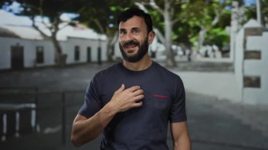 Dışarıda güneşli bir sokakta duran sakallı bir adam göğsünde bir el ile şaşkınlığını ifade ediyor.