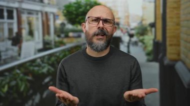 Gözlüklü ve sakallı beyaz bir adam şehir dışında arka planı bulanık ve hareketli bir şehir atmosferine sahip bir şehir ortamında yalvarıyor.