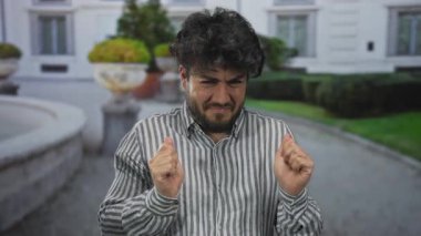 Sakallı genç adam, yeşil ve mimari geçmişi olan bir sokakta, sıradan çizgili gömleğiyle hoşnutsuzluğunu dile getiren bir jest yapıyor..