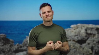 Deniz kenarına gülümseyen, yeşil tişörtlü, jest yapan, canlı bir sahil manzarasında kendine güven ve rahatlığı somutlaştıran bir adam..