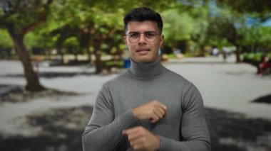 Parkta gözlüklü genç bir adam gri bir süveter giyerken jestler yapıyor, yemyeşil ağaçlar ve açık havada parlak bir çevre tarafından vurgulanıyor..