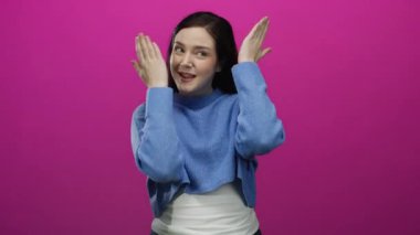 Kadın, elleri parlak pembe arka planda şakacı hareketler yapar mavi süveter giyer, hoş bir şekilde gülümser..