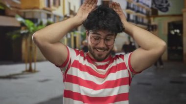 Çizgili tişörtlü, neşeli bir şekilde gülümseyen genç İspanyol bir adam. Elleri açık şehir sokaklarında neşeli bir ruh hali içinde.