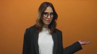 Gözlüklü genç İspanyol kadın, turuncu arka planda, spor ceket ve beyaz gömlek giyerek, özgüven ve profesyonellik yayıyor..