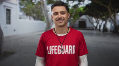 Şehir caddesinde cankurtaran tişörtü giyen genç İspanyol adam ağaçlar ve binalarla dolu canlı bir ortamda gülümsüyor..