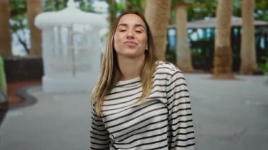 Genç sarışın kadın çizgili bir tişörtle parkta neşeyle dans ediyor, ağaçlar ve parlak güneş ışığı ile çevrili, açık havada canlı bir atmosfer yaratıyor..