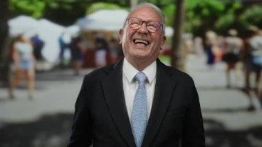 Takım elbiseli kıdemli bir adam neşeli bir şekilde neşeli bir sokakta gülümsüyor. Arka planda bulanık insanlar ve yeşillik var..