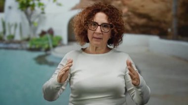 Elleriyle el kol hareketi yapan bir kadın açık havuzda yeşillik içinde.