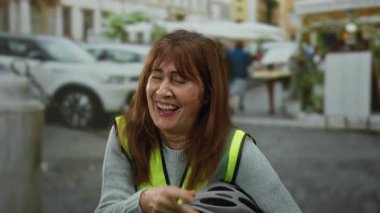 Kıdemli İspanyol kadın dışarıda gülerken şehir caddesinde bisiklet kaskı tutuyor. Canlı şehir ortamında yansıtıcı yelek giyiyor. Arabaların ve binaların geçmişi bulanık..
