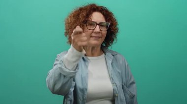 Orta yaşlı, esmer saçlı ve gözlüklü bir kadın yeşil bir arkaplanı işaret ediyor, günlük kıyafetler giyiyor ve kendine güvenen bir ifade sergiliyor..