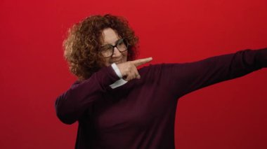 Kıvırcık saçlı ve gözlüklü, kendine güveni tam bir jest sergileyen canlı kırmızı arka planda gülümseyen bir kadın..