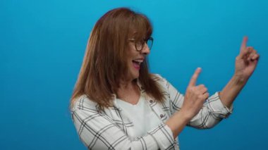 Gözlüklü yaşlı İspanyol kadın, mutlu bir şekilde gülümsüyor ve mavi arka plan duvarına işaret ediyor, beyaz ekose gömlek giyiyor..