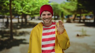 Parkta neşeli bir şekilde gülümseyen İspanyol bir adam, kırmızı bir bere ve sarı yağmurluk giyiyor, arka planda yemyeşil ağaçlar var..