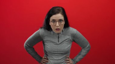 Esmer kadın, koyu kırmızı arka planda, gözlüklü ve gri kazaklı bir kadına şaşkınlık ve şaşkınlık içinde bakıyor..