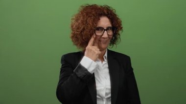 Gözlüklü bir kadın titiz bir jest yapıyor, düz yeşil bir arka plana karşı duruyor, resmi bir takım elbise giyiyor, farkındalık ve ciddiyet duygusunu gösteriyor..