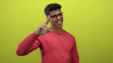 Gözlüklü genç adam pembe kazak giyiyor. Parlak sarı arka plana karşı neşeli poz veriyor. Kendine güveni, mutluluğu ve modern tarzı gösteriyor.