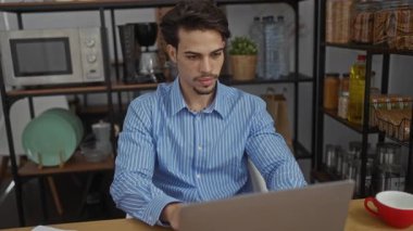 Çizgili gömlekli genç bir adam ofisteki bir masada oturuyor, bir dizüstü bilgisayarı kapatıyor ve kollarını açıyor, mutfak raflarıyla çevrili, çeşitli eşyalar ve bir bitki ile..