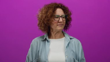 Elleri parlak pembe arka plana uzanmış, gözlük takan, kıvırcık saçlı ve tarafsız ifadeli mavi gömlekli bir kadın.