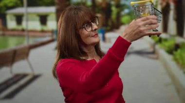 Kırmızı kazaklı kadın açık hava parkında arka planı bulanık mini alışveriş arabası tutuyor. Eğlenceyi ve yaşam tarzını vurgulayan neşeli bir an sergiliyor..