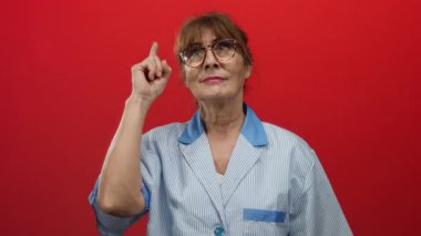 Temizlik üniformalı kıdemli İspanyol kadın canlı kırmızı bir arka plana karşı düşünceli bir ifadeyle işaret ediyor..