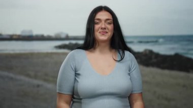 Uzun saçlı, gülümseyen ve kumsaldaki kameraya bakan bulutlu bir günde arka planda deniz olan genç bir kadın kendinden emin ve kıvrımlı vücudunu sergiliyor..