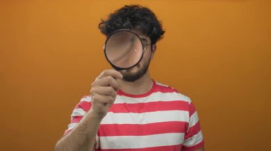 Elinde büyüteç tutan, turuncu arka planda çizgili gömlek giyen genç bir adam.