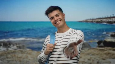 Çizgili gömlekli genç adam, elini kameraya doğru uzatıyordu. Arka planda turkuaz bir deniz vardı. Dostane bir yaz anını yakalıyordu..