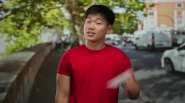 Elinde öğrenci tabelası tutan genç adam şehir caddesinde arabaların ve ağaçların arasında günlük kırmızı gömleğiyle canlı bir çevrede şehir yaşamını gözler önüne seriyor..