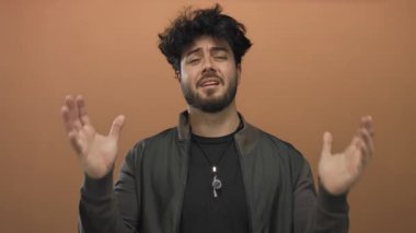 Genç sakallı İspanyol adam, portakal arka planına uzanan ellerini uzatarak kafa karışıklığı ve duygularını ifade ediyor..