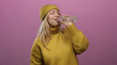 Sarı süveter ve bereyle canlı pembe bir duvara yaslanmış su şişesinden hoşlanan bir kadın, su ve konfor yayıyor..