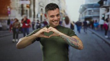 Dövmeleri olan genç bir adam şehir sokaklarında kalp şekli oluşturur. Arka planda bulanık yayalar şehir ortamında sevgi ve bağlantı hissi taşır.