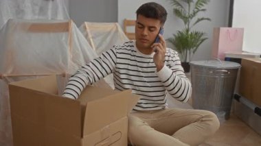 Yeni evdeki kutuları açarken telefonla konuşan genç bir adam. Etrafı mobilyalarla çevrili. Yeni bir yaşam alanına geçişi gösteriyor..