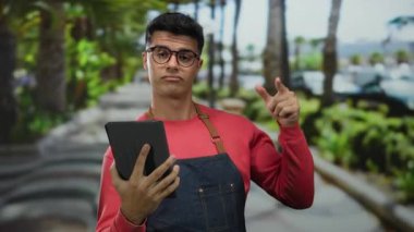 Gözlüklü genç adam dışarıda, yemyeşil bir park ortamında tablet kullanıyor, günlük kırmızı kıyafetler ve önlük giyiyor, odaklanmış ve dokunmatik ekran cihazıyla meşgul görünüyor..