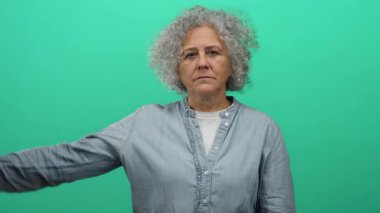 Gri saçlı yaşlı bir kadın, izole edilmiş yeşil arka plan duvarına karşı güçlü bir jest yapıyor..