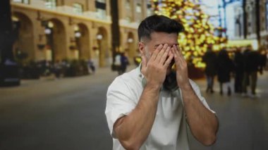 Boynunda kulaklıklarla gülümseyen genç bir adam aydınlatmalarla süslenmiş bir şehir caddesinde ve bir noel ağacıyla açık hava Noel atmosferinin tadını çıkarıyor..