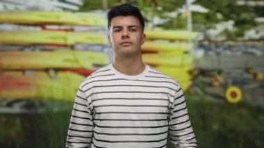 Sahilde askeriyeye selam veren genç bir adam, arka planda kanoları olan, çizgili gömlek giyen, kendinden emin görünen ve deniz tarafından bestelenmiş, açık hava macerasını ve saygıyı temsil eden biri..