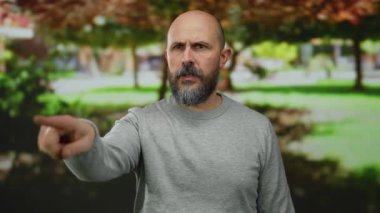 Sakallı bir adam park ortamını işaret ediyor. Yüzü bulanık, çevreye odaklanıyor. Gri süveter giyiyor..