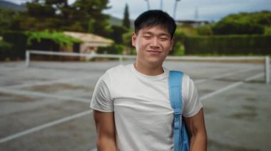 Çinli bir öğrenci açık tenis kortunda, mavi bir sırt çantasıyla gülümsüyor, gençlik enerjisini ve spor ortamını somutlaştırıyor..