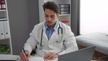 Klinikte steteskop takan genç bir adam, tıbbi dosyalar ve iç sağlık hizmetlerindeki dizüstü bilgisayarlarla çevrili bir masadaki belgelere yazıyor..