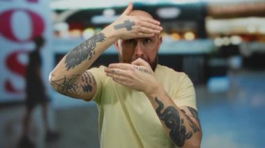 Dövmeleri olan bir adam neşeli bir şekilde kapalı bir alışveriş merkezi ortamında yüzünü örtüyor. Duygularını ve tarzını açık seçik ifade ediyor..