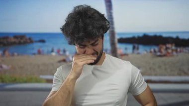 Sahilde sakallı genç İspanyol bir adam hapşırıyor, arka planda palmiye ağacı ve deniz görünüyor, açık deniz sahili anını yakalıyor..